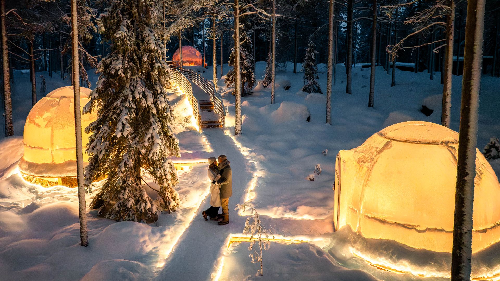 Couple walking near illuminated Aurora igloos at Invisible Forest Lodge.