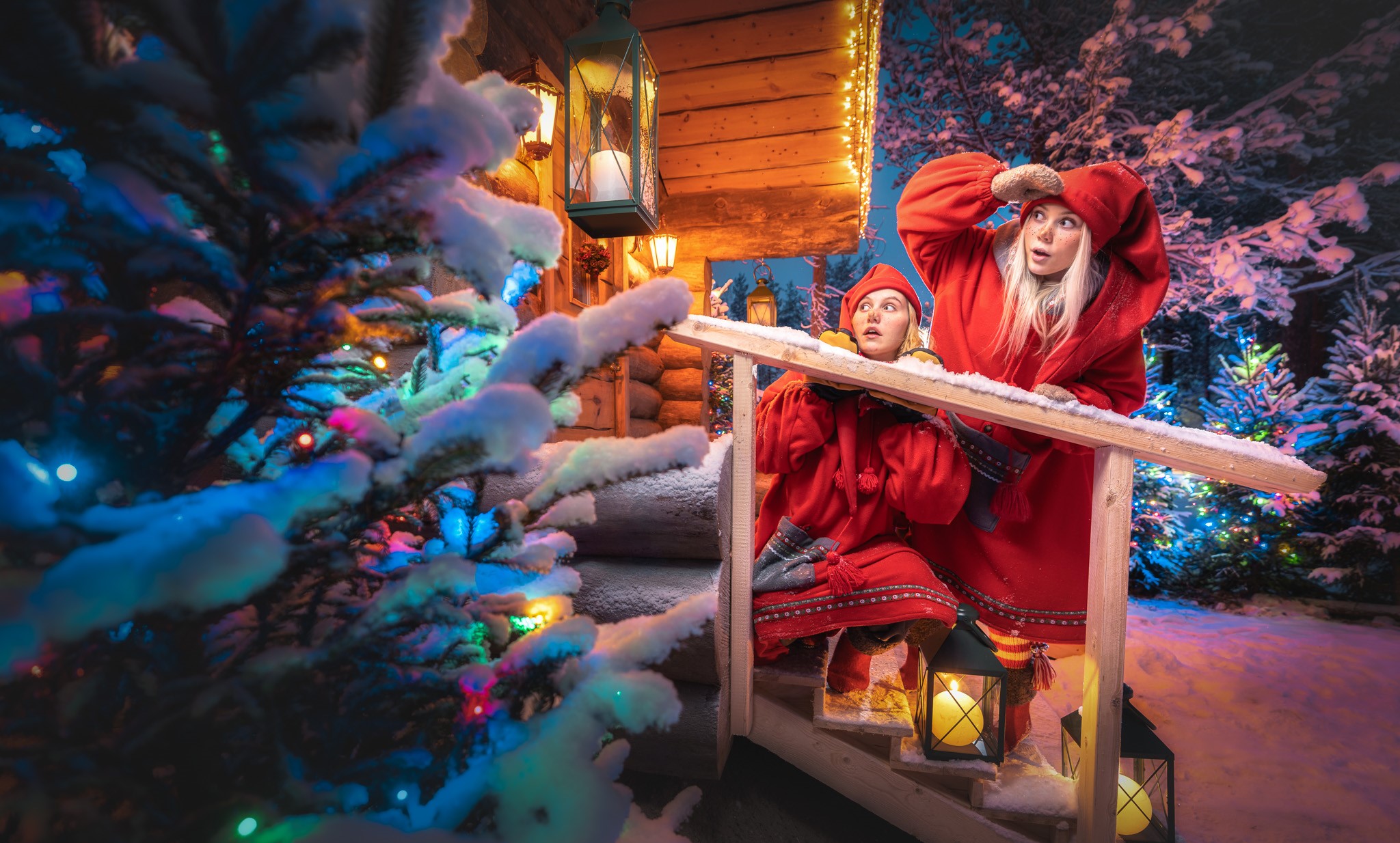 two elves in front of the log cabin with christmas tree in the front of the photo.