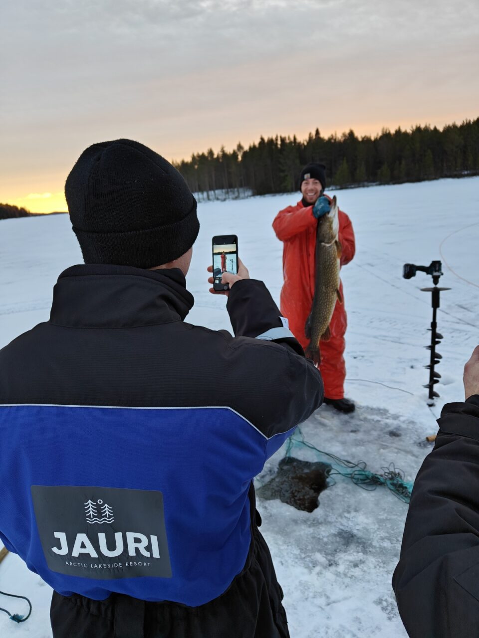 Person taking a photo of a man holding a fish on frozen lake.