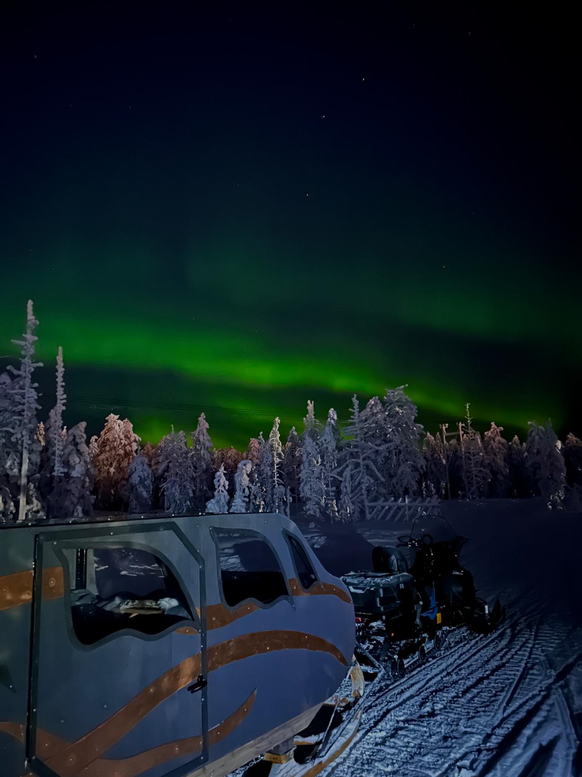 Auroras illuminate snowy landscape at night with a sledge pulled by a snowmobile.