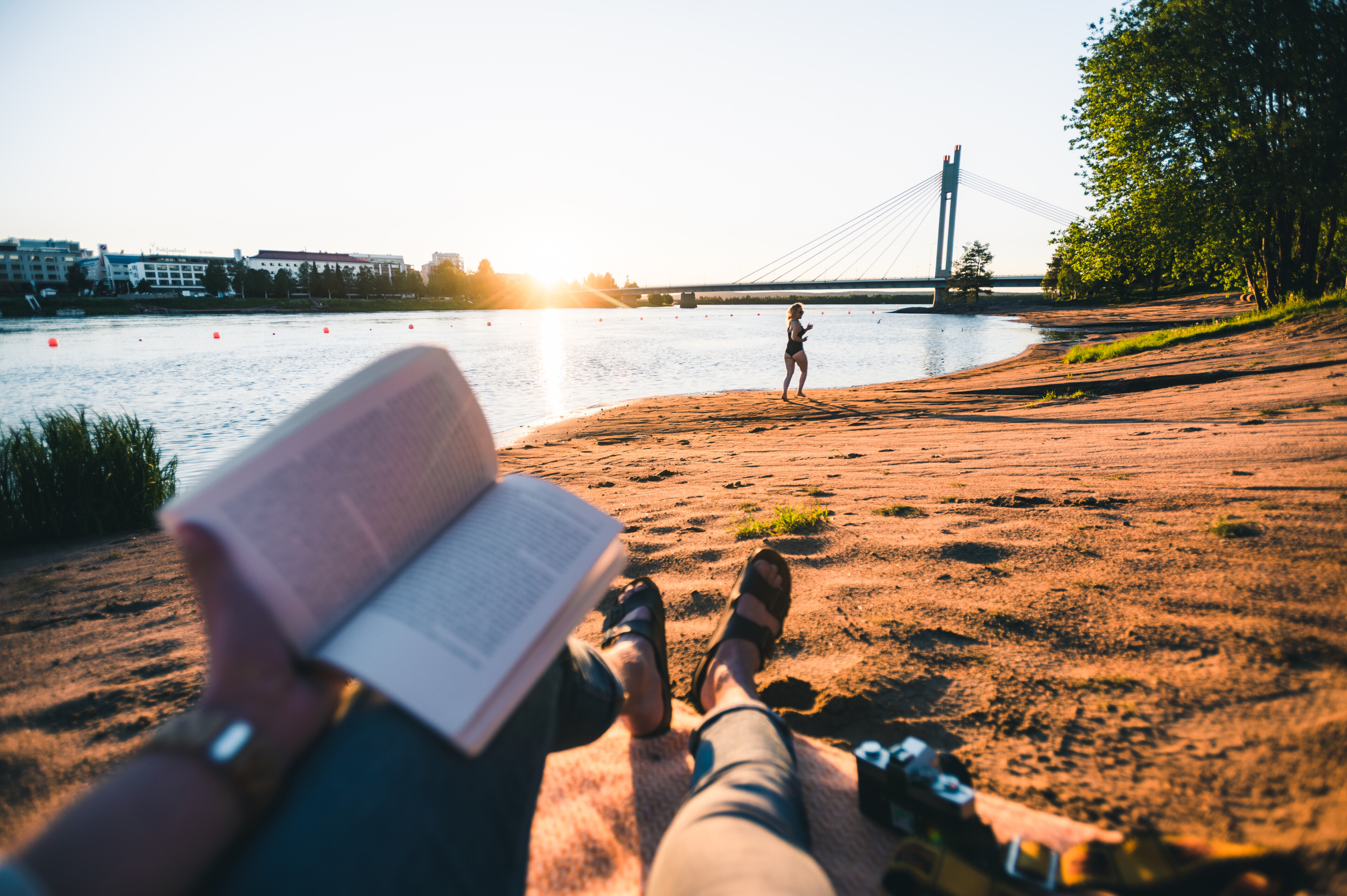 Ihminen istuu rannalla kesällä keskiyön auringossa taustalla Jätkänkynttilän silta, Rovaniemellä, Lapissa.